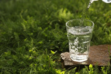 Pouring fresh water from bottle into glass on stone in green grass outdoors. Space for text