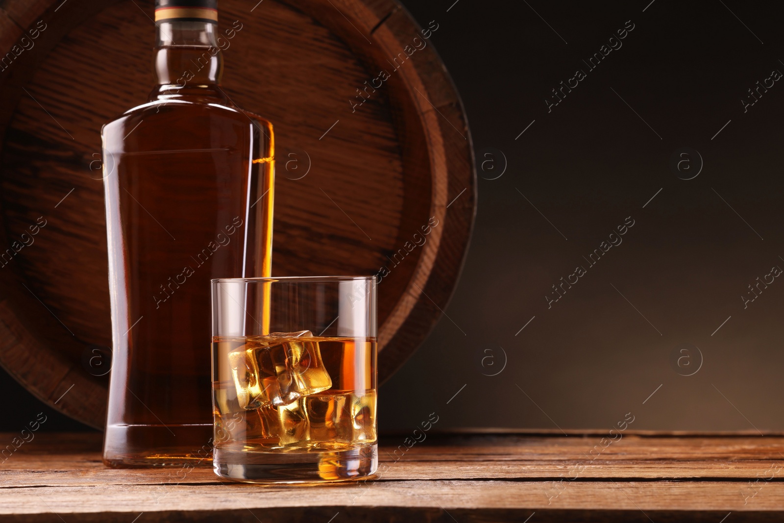 Photo of Whiskey with ice cubes in glass, bottle and barrel on wooden table against black background, space for text