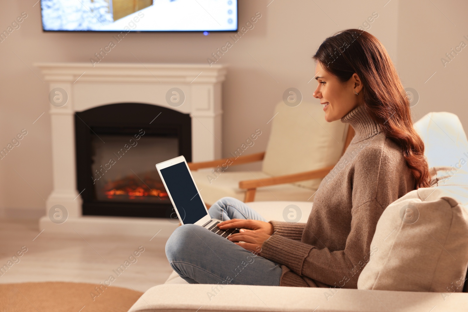 Photo of Young woman with laptop on sofa near fireplace at home