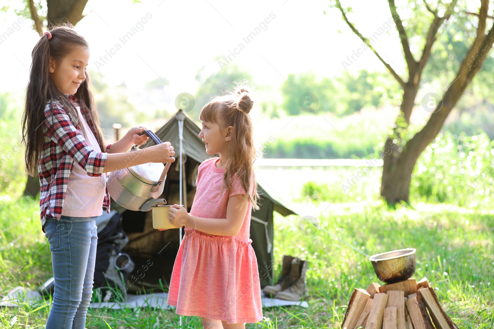 Photo of Little girls with mug and kettle near tent outdoors. Summer camp