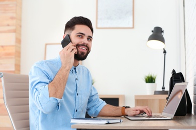 Handsome young man talking on phone while working at table with laptop in home office