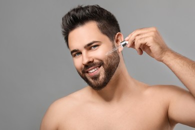 Handsome man applying cosmetic serum onto face on grey background, space for text