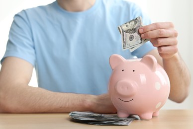 Photo of Financial savings. Man putting dollar banknote into piggy bank at wooden table, closeup