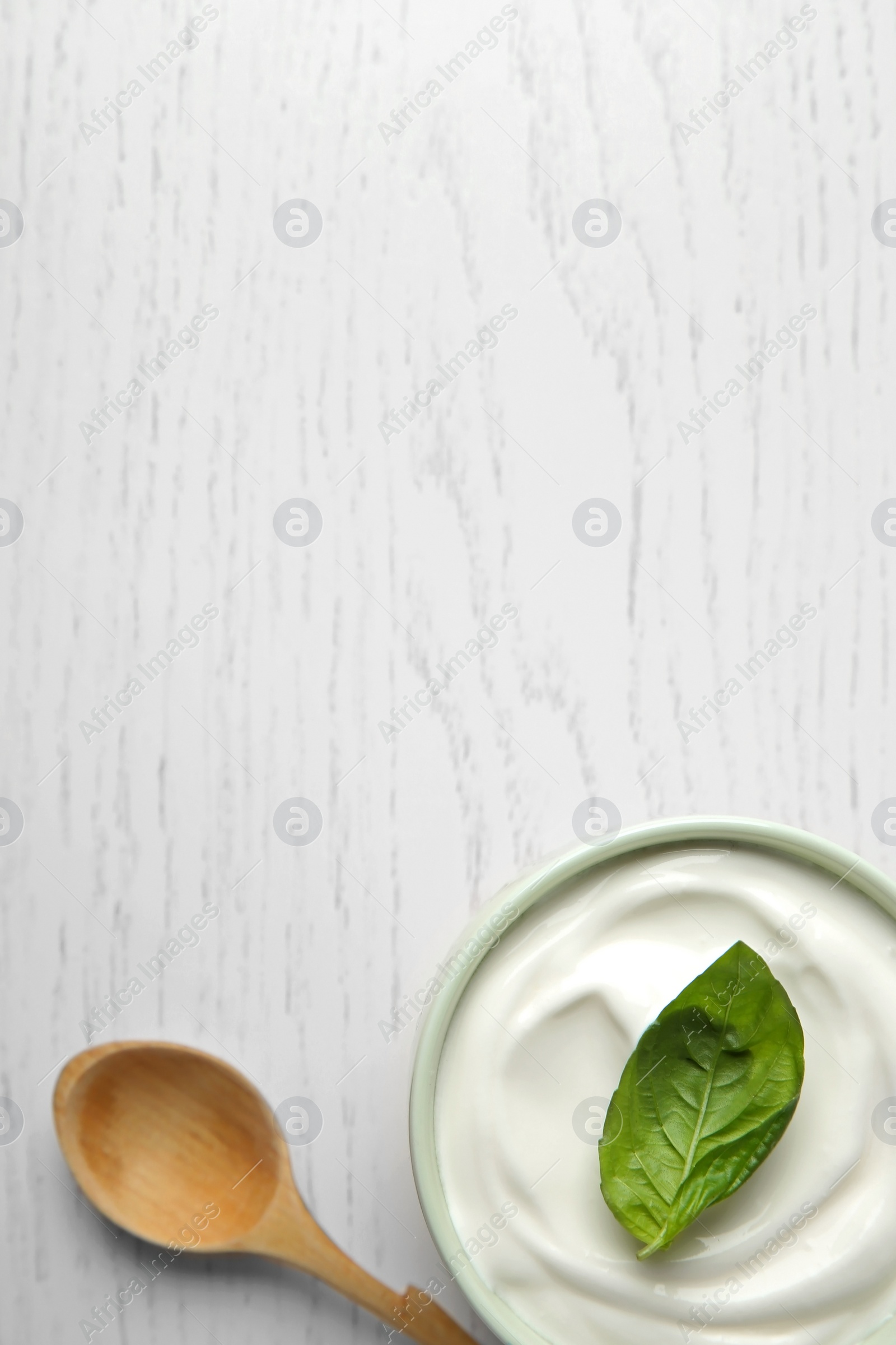 Photo of Bowl of fresh sour cream with basil and spoon on white wooden table, flat lay. Space for text