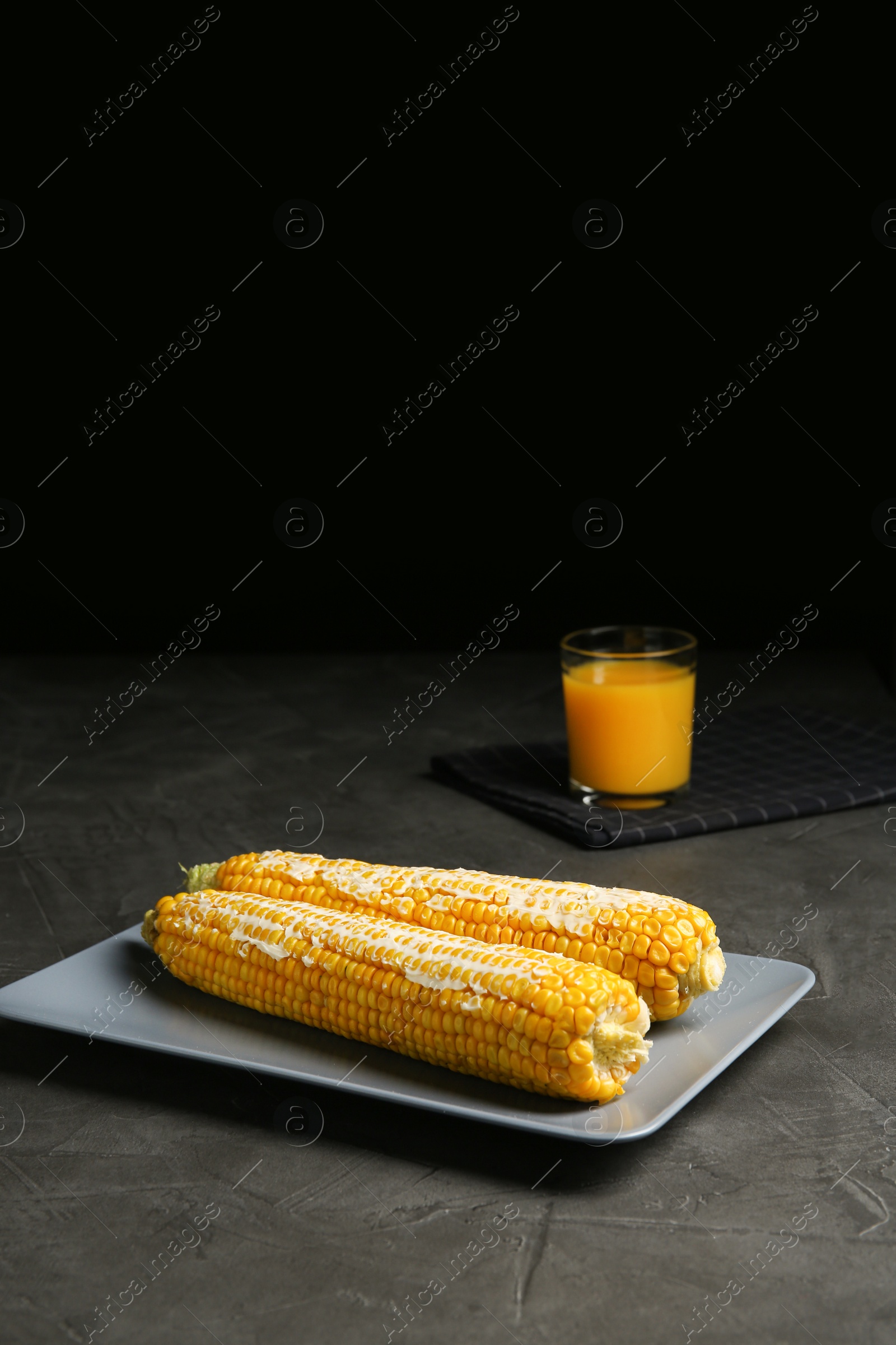 Photo of Delicious boiled corn cobs with butter on gray table against black background. Space for text