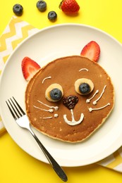 Photo of Creative serving for kids. Plate with cute cat made of pancakes, berries, cream, banana and chocolate paste on yellow background, flat lay