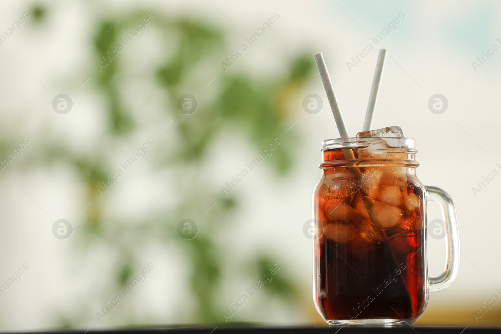 Photo of Mason jar of cola with ice on table against blurred background. Space for text