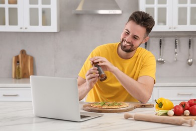Photo of Happy man adding pepper on pizza while watching cooking online course in kitchen. Time for hobby