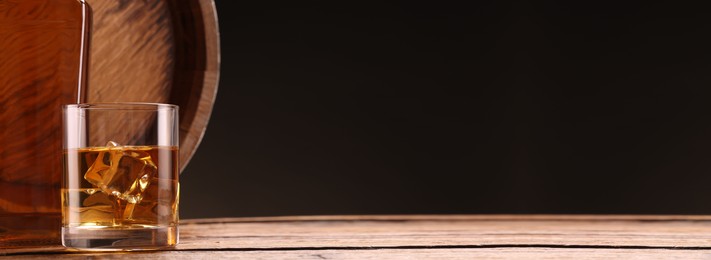 Whiskey with ice cubes in glass, bottle and barrel on wooden table against black background, space for text. Banner design