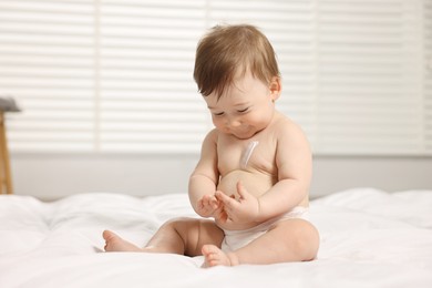 Photo of Cute little baby with moisturizing cream onto body sitting on bed at home