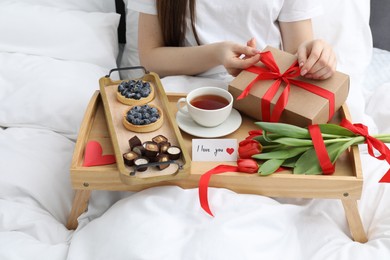 Photo of Tasty breakfast served in bed. Woman with gift box, desserts, tea, flowers and I Love You card at home, closeup