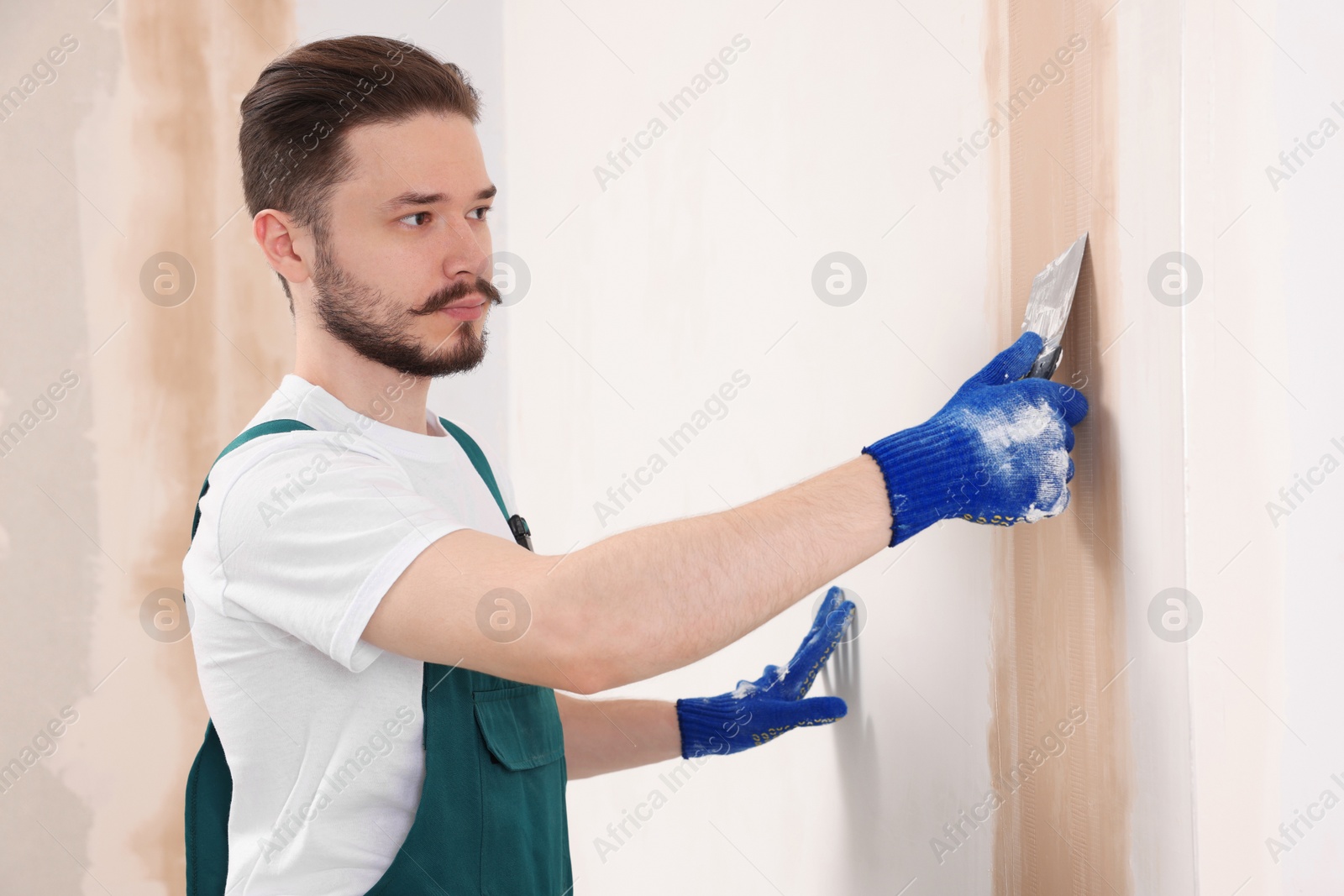 Photo of Worker in uniform plastering wall with putty knife indoors