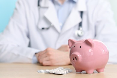 Doctor with pills at wooden table, focus on piggy bank