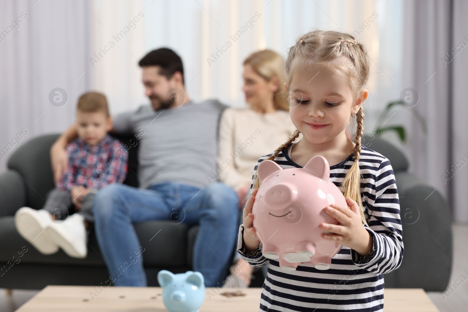 Photo of Family budget. Little girl with piggy bank, her parents and brother at home, selective focus