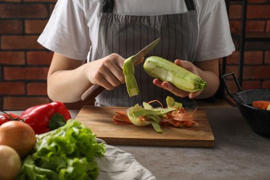 Woman peeling fresh zucchini with knife at grey table indoors, closeup