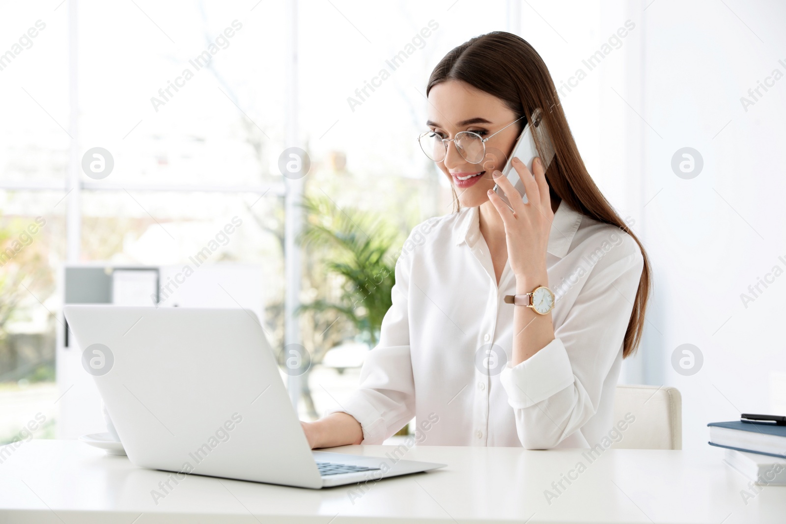 Photo of Young businesswoman talking on phone while using laptop at table in office