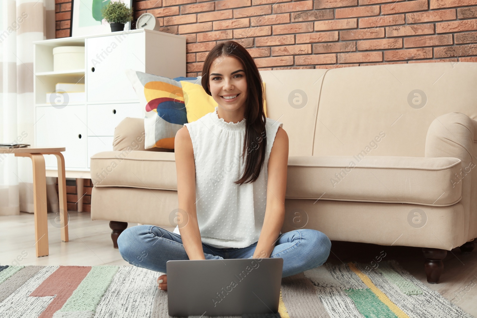 Photo of Young woman with modern laptop sitting on floor near sofa at home