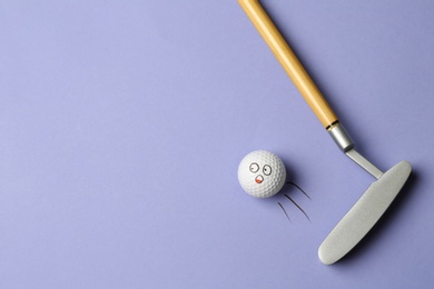 Photo of Golf ball with funny face flying away from club on lilac background - creative image. Top view
