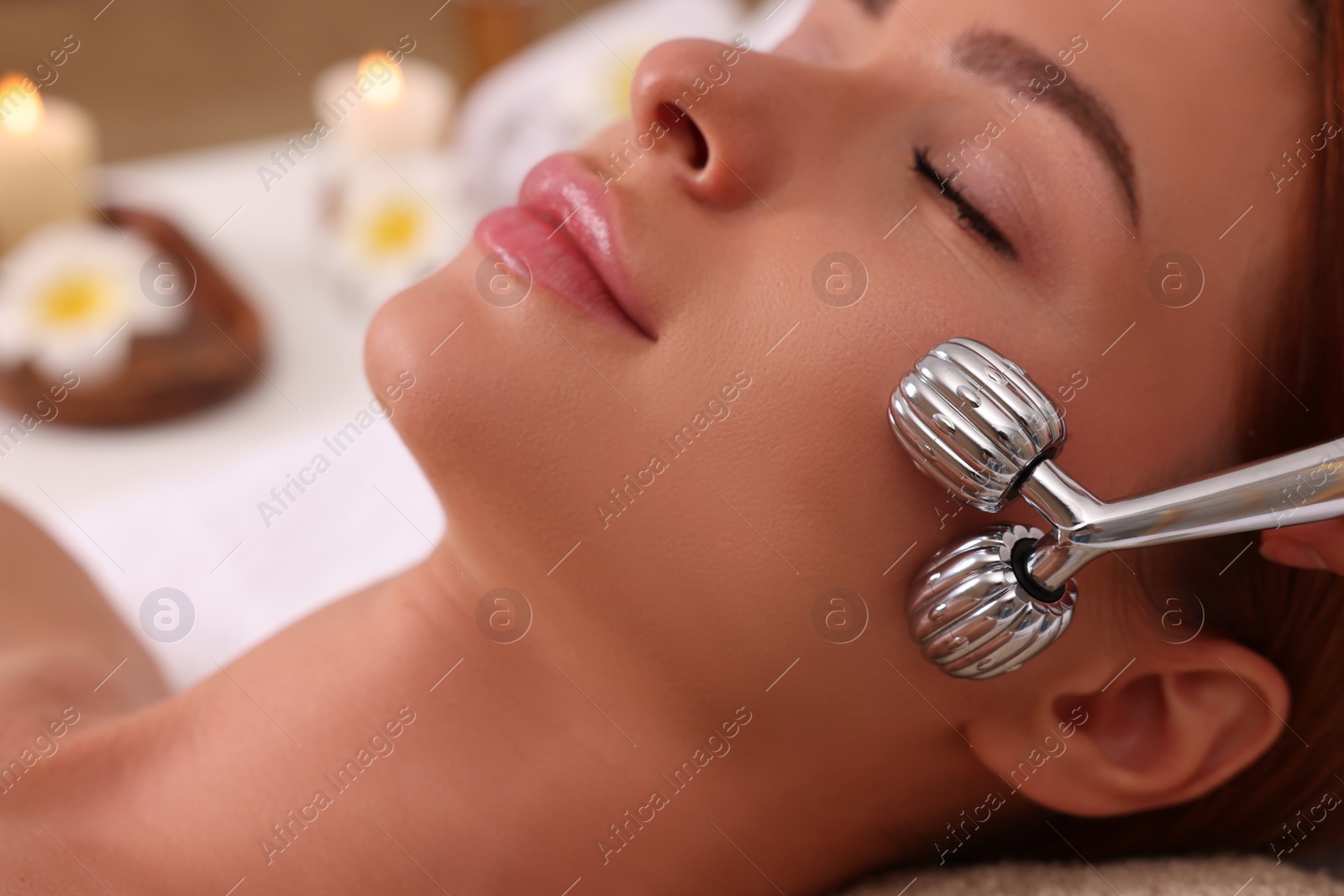 Photo of Young woman receiving facial massage with metal roller in beauty salon, closeup