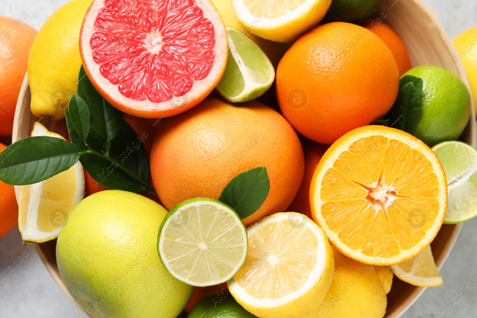 Photo of Different fresh citrus fruits and leaves in bowl on light table, top view