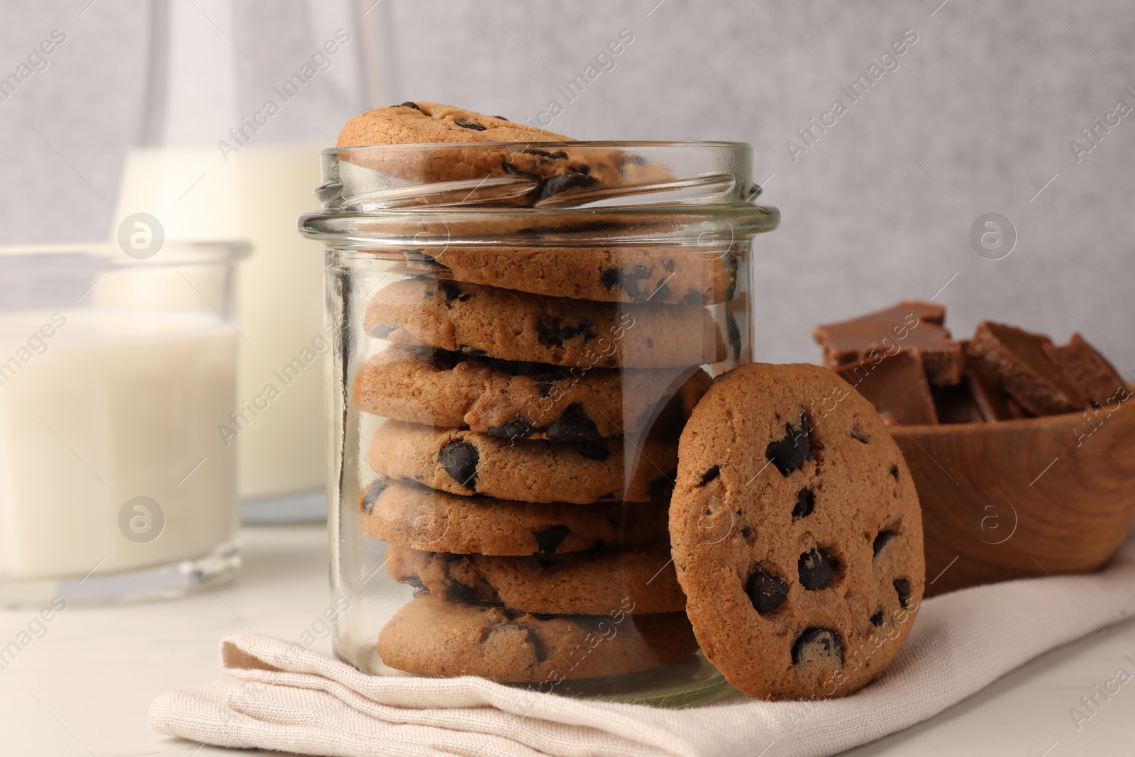 Photo of Glass jar with delicious chocolate chip cookies and milk on white marble table, closeup