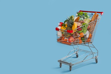 Photo of Shopping cart full of groceries on light blue background. Space for text
