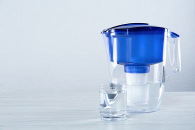 Photo of Filter jug and glass with purified water on white table against light background. Space for text