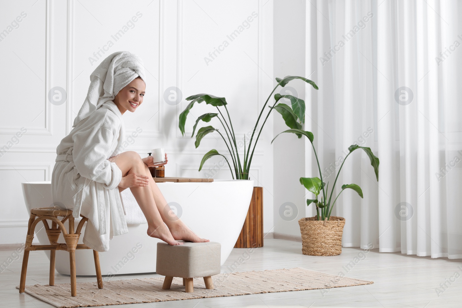 Photo of Beautiful young woman applying body cream onto legs in bathroom, space for text