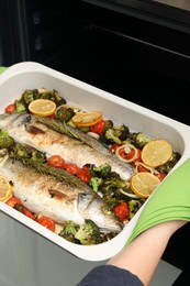 Woman taking baking dish with delicious fish and vegetables from oven, closeup