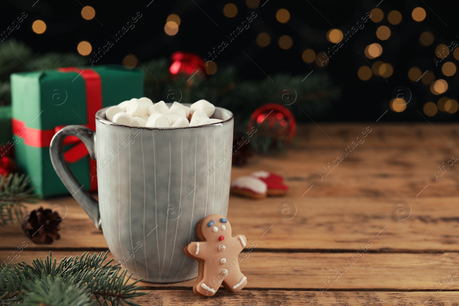 Photo of Delicious hot chocolate with marshmallows and gingerbread cookie on wooden table against blurred lights, space for text