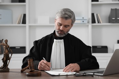 Photo of Judge with gavel and laptop writing in papers at wooden table in office