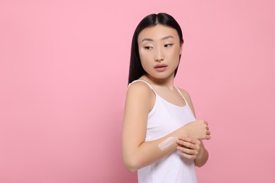 Photo of Beautiful young Asian woman applying body cream onto arm on pink background, space for text