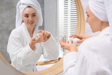 Beautiful woman in white robe applying cream near mirror indoors