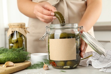 Woman taking pickled cucumber from jar at table in kitchen, closeup view. Space for text