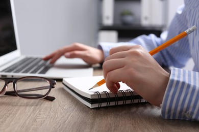 Photo of Woman with notebook working on laptop at wooden table in office, closeup