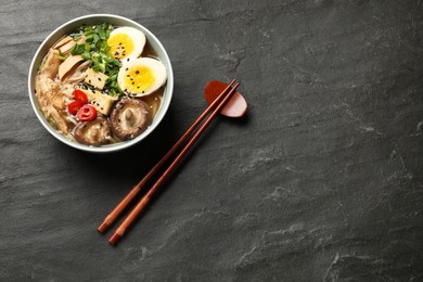 Noodle soup. Bowl of delicious ramen, ingredients and chopsticks on black table, flat lay. Space for text