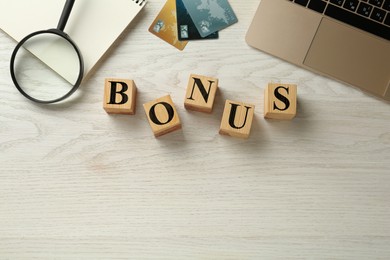 Image of Word Bonus made of wooden cubes with letters, credit cards, laptop, magnifying glass and notebook on white wooden tablet, flat lay