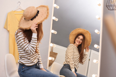 Beautiful young woman wearing hat looking at herself in large mirror indoors