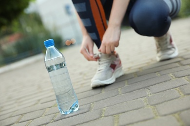 Closeup view of sportswoman lacing sneakers outdoors, focus on bottle with water