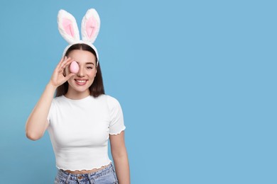 Photo of Happy woman in bunny ears headband holding painted Easter egg on turquoise background. Space for text