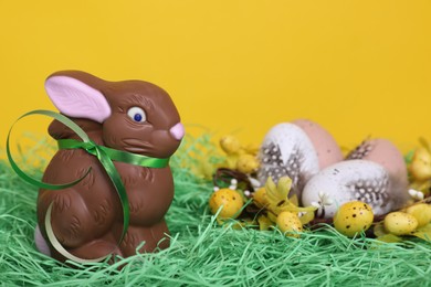 Easter celebration. Cute chocolate bunny and different eggs with feathers on grass against yellow background