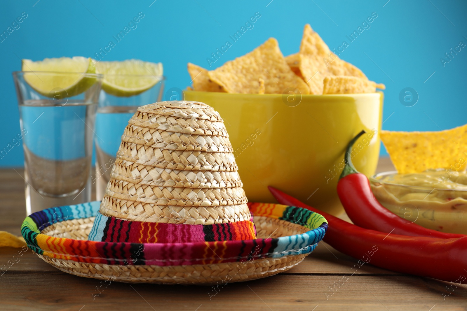 Photo of Mexican sombrero hat, tequila, chili peppers, nachos chips and guacamole on wooden table against light blue background