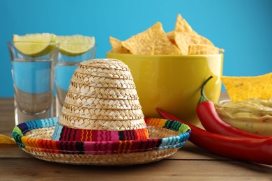 Mexican sombrero hat, tequila, chili peppers, nachos chips and guacamole on wooden table against light blue background