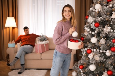 Photo of Young woman decorating Christmas tree while her boyfriend resting on sofa at home