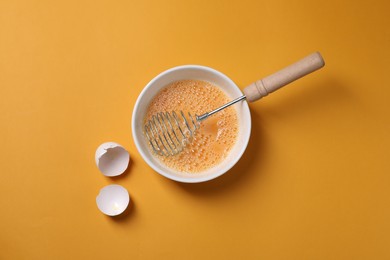 Photo of Beaten eggs, metal whisk in bowl and shells on orange background, flat lay