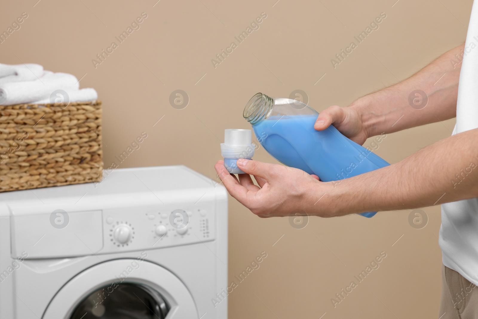 Photo of Man pouring fabric softener from bottle into cap near washing machine indoors, closeup