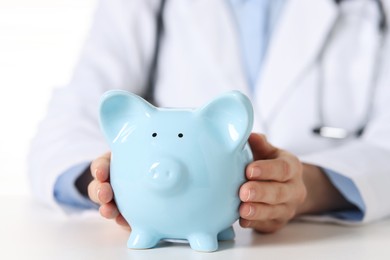 Photo of Doctor with piggy bank at white table, closeup