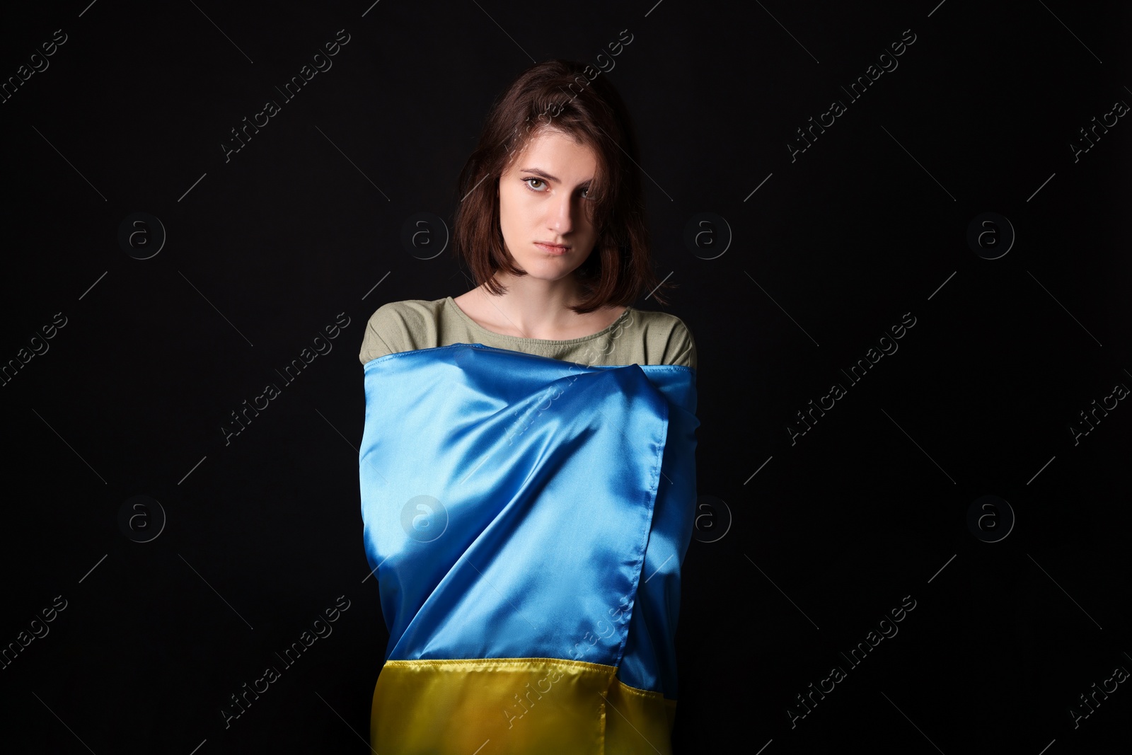 Photo of Sad woman wrapped in Ukrainian flag on black background
