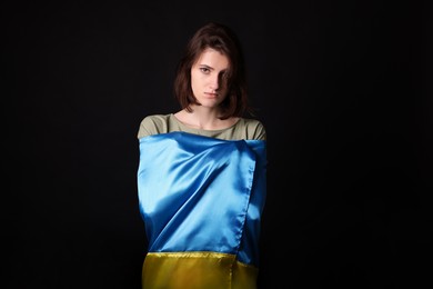 Photo of Sad woman wrapped in Ukrainian flag on black background