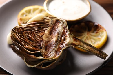 Tasty grilled artichoke, slices of lemon and sauce on table, closeup